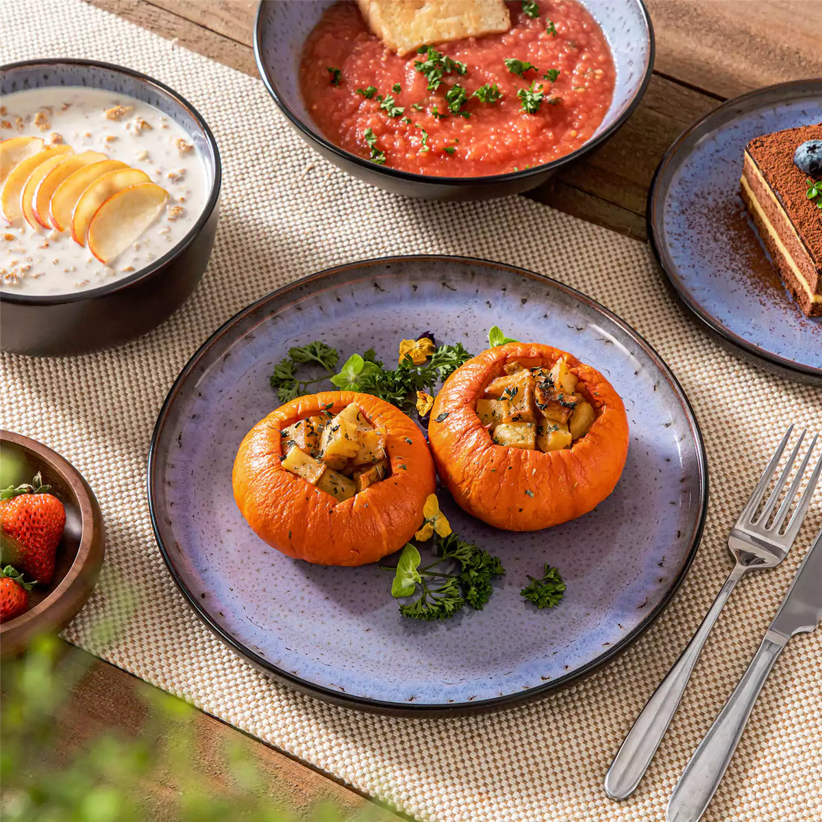 Purple stoneware set for one, with matte black exterior and glossy violet glaze, featuring roasted pumpkin, soup, and oats on a wooden table with linen placemats.