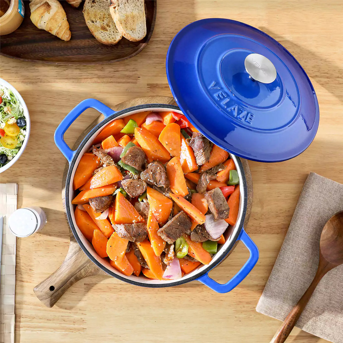6qt blue enameled cast iron round Dutch oven with lid on beige wooden table, serving tender beef short ribs, accompanied by a salad bowl.