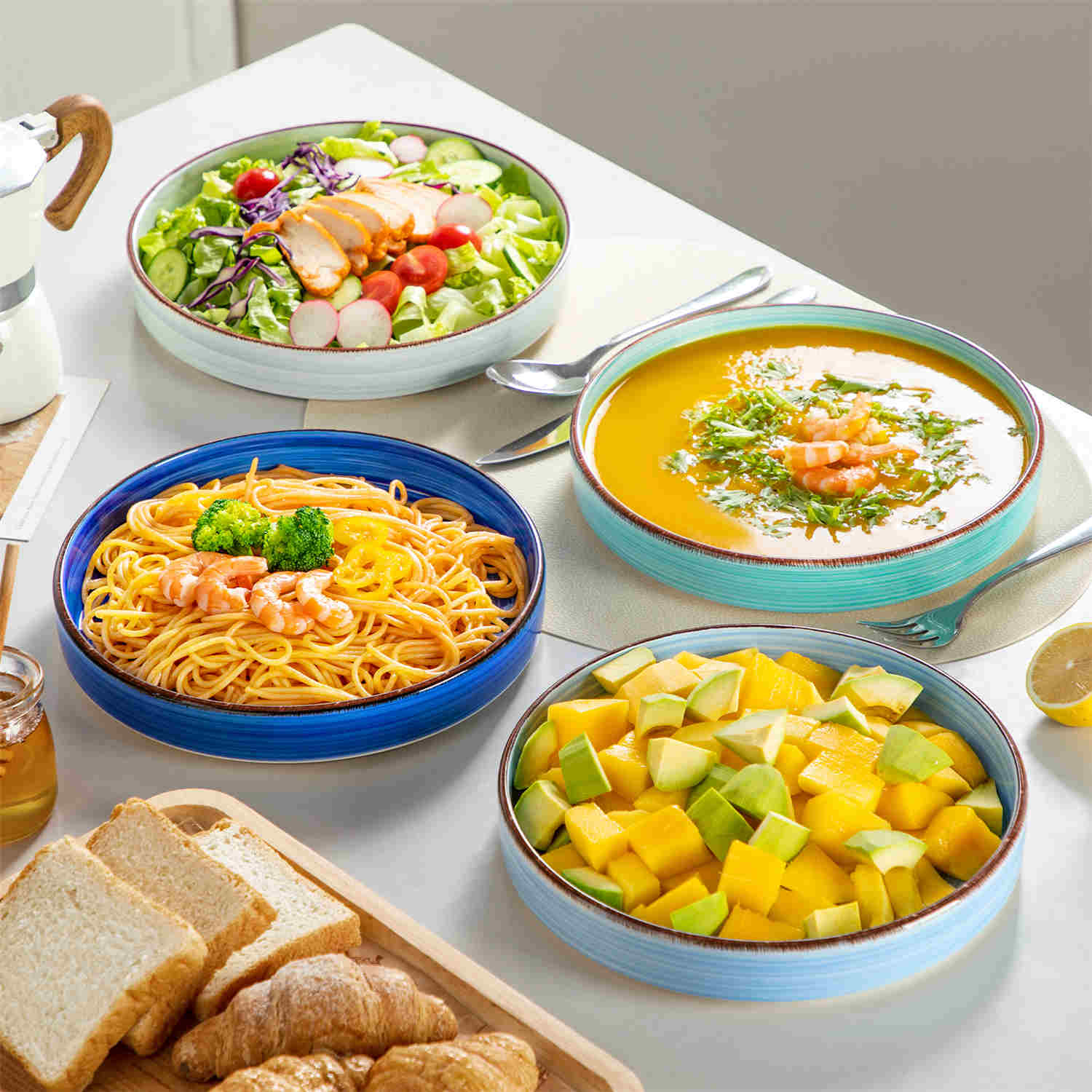 Bonita straight-edge pasta bowls set of 4 on a white table - filled with spaghetti - fruit salad - and mango - accompanied by a small honey jar - ibrant stoneware in ocean blues