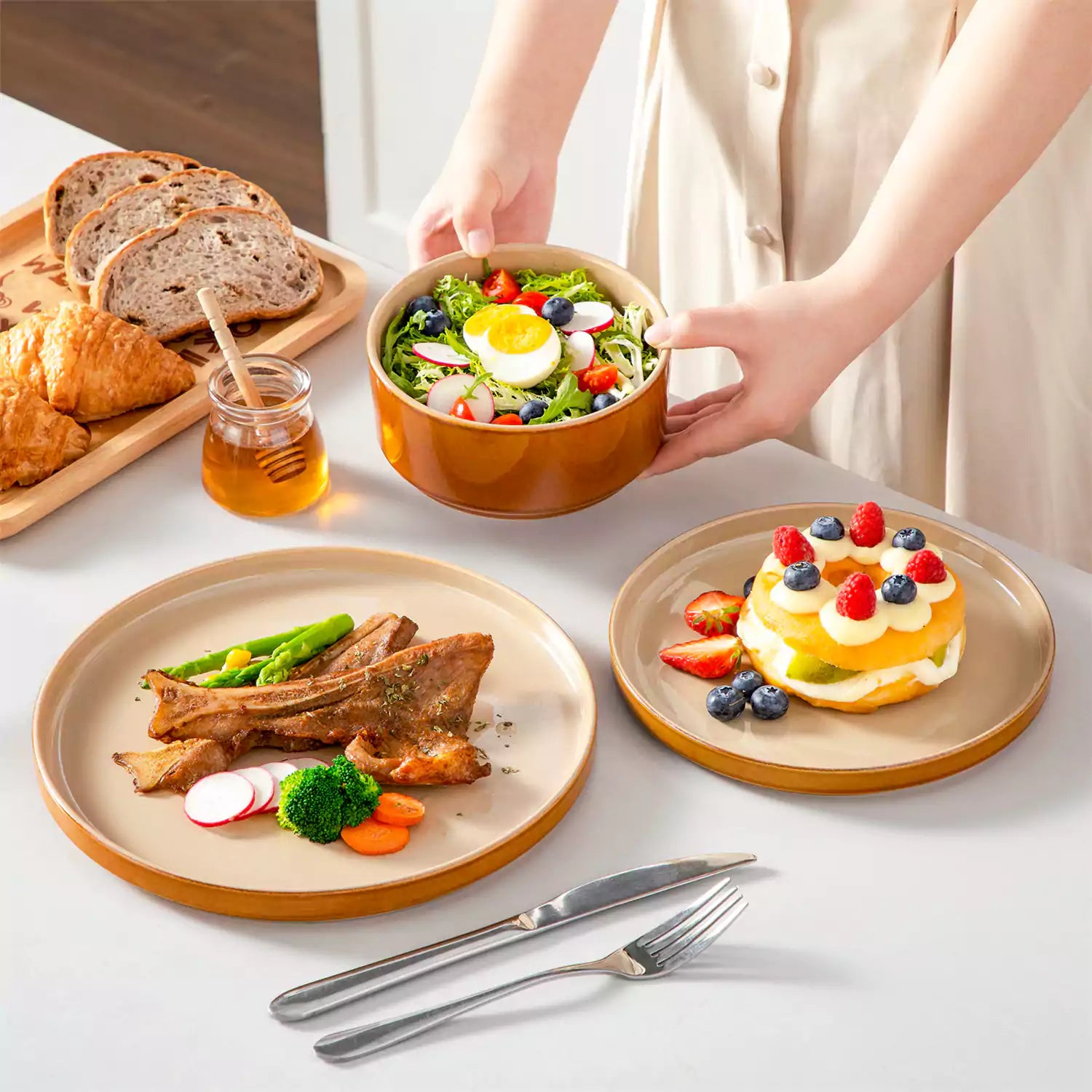 A rustic breakfast spread featuring a plate of meat with vegetables - waffles topped with fruit - and assorted pastries on a white table - complemented by caramel stoneware dinnerware