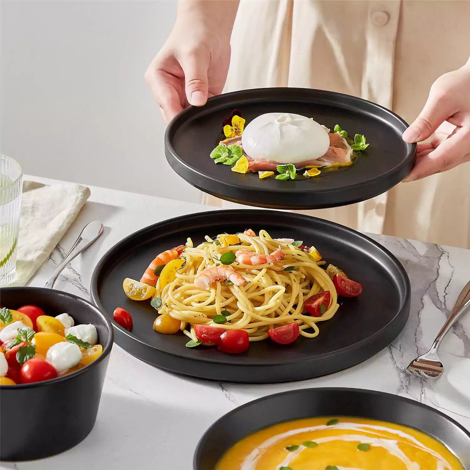 Modern black stoneware bowl and plate set displayed on a marble countertop - with a fresh salad blurred in the background - vancasso
