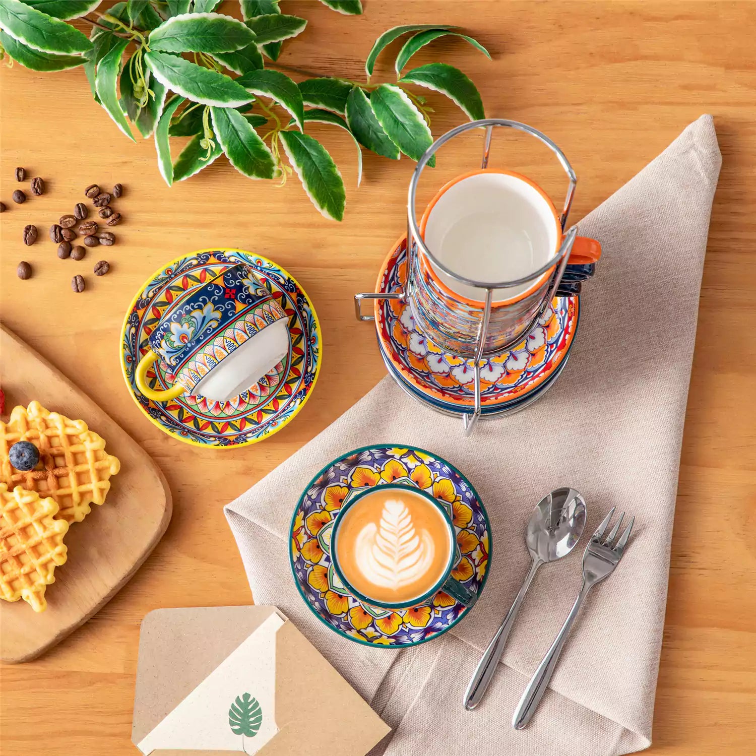 Simi 12 piece porcelain cafe set with 5oz flat white cups and saucers in vibrant boho chic floral patterns on a black metal storage rack with a magazine and napkin on a wooden table