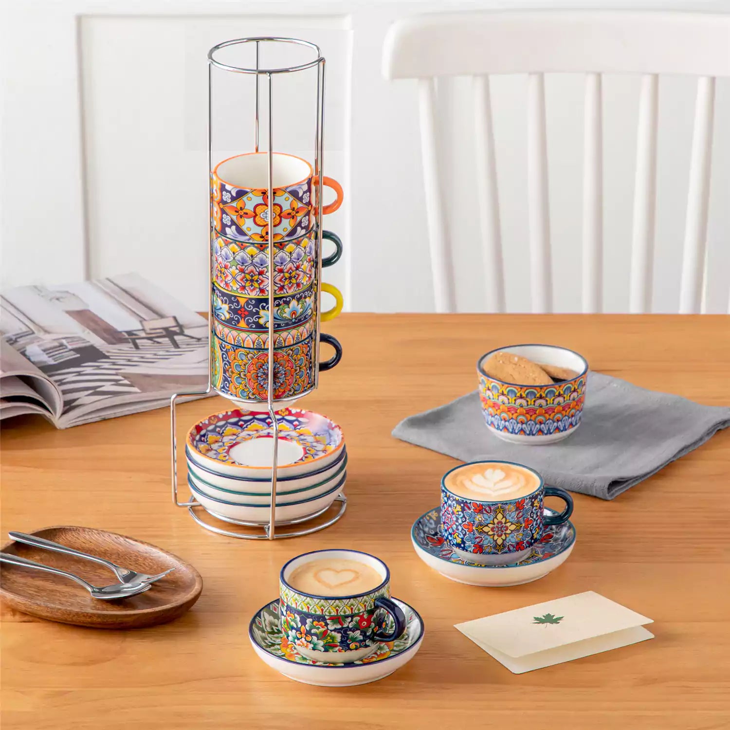 Simi 12 piece porcelain cafe set with 5oz flat white cups and saucers in vibrant boho chic floral patterns on a black metal storage rack with a magazine and napkin on a wooden table