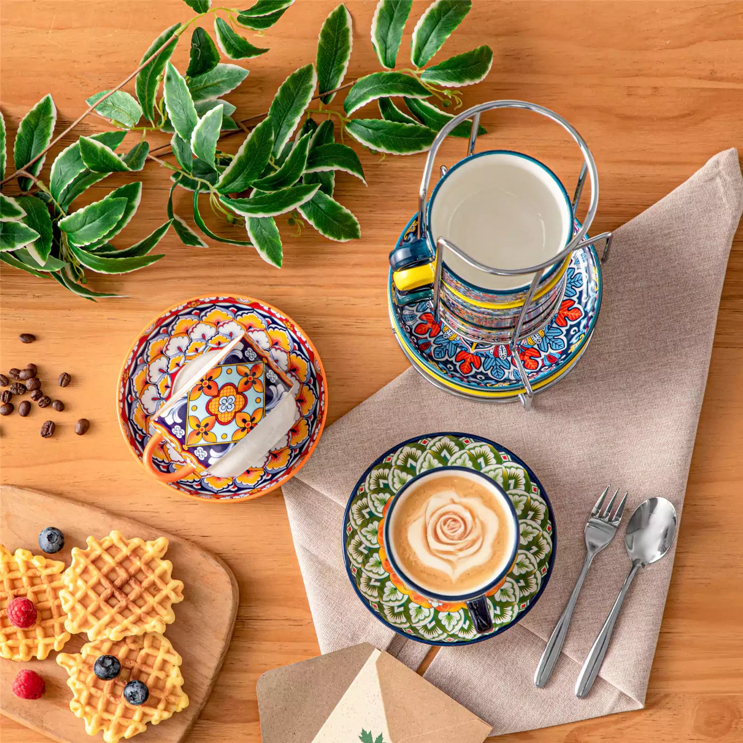 Simi 12 piece porcelain cafe set with 9oz flat white cups and saucers in vibrant boho chic floral patterns on a black metal storage rack with a magazine and napkin on a wooden table